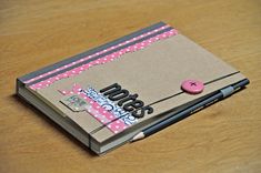 a notebook with a pink button on it sitting on top of a wooden table next to a pen