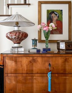 a wooden dresser topped with a vase filled with pink flowers next to a painting and lamp