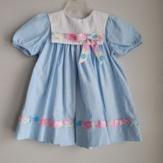 "This adorable vintage dress by Rare Editions is perfect for spring! The dress is made of 100% cotton blue chambray denim. There are short sleeves and buttons at back of neck. There is a square white eyelet collar with floral grosgrain ribbon trim and bow. The trim is mimicked at the hem. Size is: 3t Length: 19.5\" chest: 14\"" Retro Spring Playtime Dresses, Blue Retro Cotton Dresses, Retro Blue Cotton Dresses, Vintage Cotton Playtime Dress, Cute Fitted Cotton Vintage Dress, Spring Blue Vintage Dress, Blue Cotton Vintage Dress For Summer, Spring Blue Cotton Vintage Dress, Blue Cotton Vintage Summer Dress