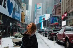 a woman standing in the middle of a snow covered street with cars and buildings behind her