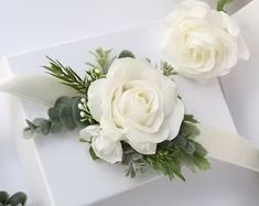 two white roses and greenery sit on top of a white gift box with ribbon