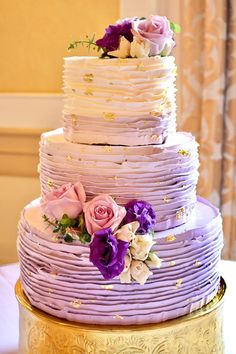 a three tiered cake with purple and white flowers on the top is sitting on a gold plate