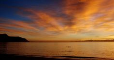 the sun is setting over the ocean with clouds in the sky and mountains in the distance