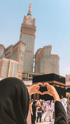 a person making a heart shape with their hands in front of a clock tower and other buildings