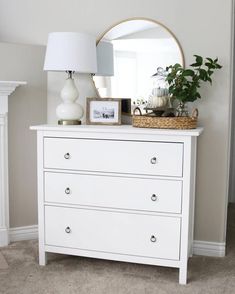 a white dresser with a mirror, lamp and plants on it in a room that has carpeted flooring