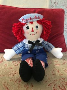 a raggedy - faced doll sits on a red pillow in front of a white wall