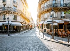 a cobblestone street with tables and chairs in the middle, surrounded by tall buildings