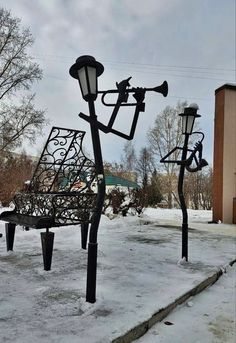 a park bench sitting next to two street lamps on top of snow covered ground with buildings in the background