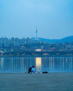 two people are sitting on the beach watching the city lights shine in the water behind them