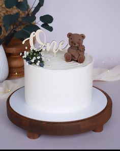 a white cake topped with a teddy bear and the word one on top is sitting on a wooden stand