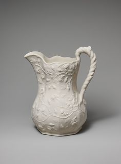 a white vase with an intricate design on the bottom and handle is shown against a gray background