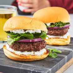 two hamburgers with lettuce, onions and cheese on a slate board next to a beer