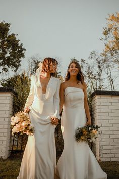 two women in white dresses standing next to each other holding hands and smiling at the camera