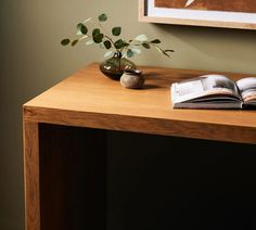 an open book on a wooden table next to a vase with leaves and a potted plant