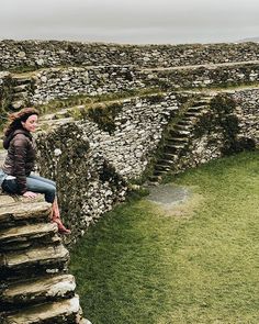 a woman sitting on top of a stone wall next to a lush green field covered in grass