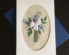 an embroidered card with flowers and leaves is displayed on top of a white envelope that sits on a wooden table