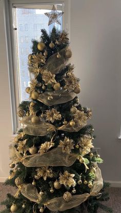 a christmas tree decorated with gold ornaments and ribbons in front of a window, on carpeted floor