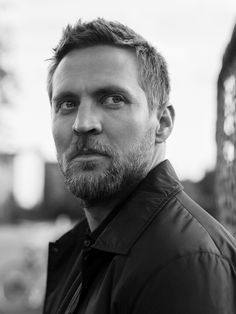 a black and white photo of a man with a beard looking at the camera while standing in front of a building