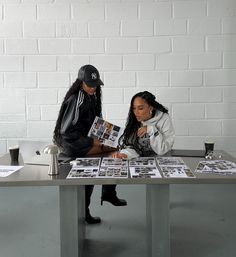 two women sitting at a table with papers and magazines on it, one reading the paper