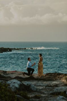 a man kneeling down next to a woman on top of a rocky cliff near the ocean