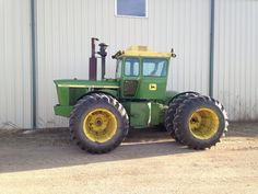 a large green tractor parked in front of a white building with yellow rims on it's tires