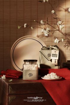 a table topped with plates and containers filled with rice next to a red cloth covered box