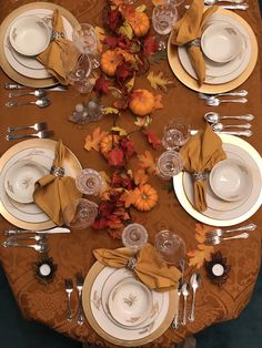 the table is set for thanksgiving dinner with white plates and silverware, gold napkins and autumn leaves