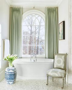 a large white bath tub sitting under a window next to a chair and vase with flowers