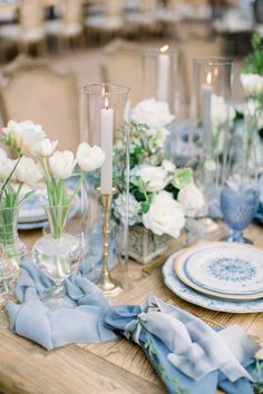 a wooden table topped with plates and vases filled with white flowers next to candles