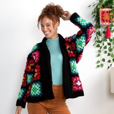 a woman wearing a colorful crochet cardigan standing next to a potted plant