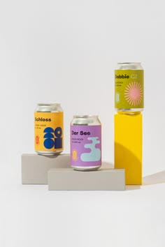 three cans of soda sitting on top of a white block next to a yellow box