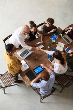 a group of people sitting around a wooden table with laptops on top of it