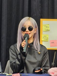 a woman with pink hair and sunglasses holding a microphone in front of a sign that reads,