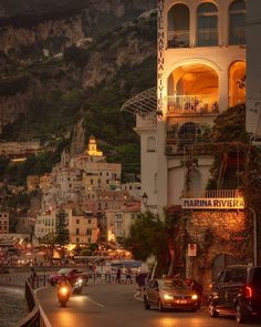 cars are driving down the road in front of some buildings and cliffs at night time