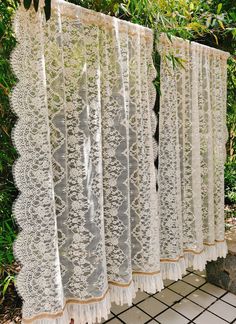 lace curtains are hanging on the side of a wall in front of some bushes and trees