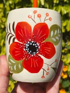 a hand holding a painted cup in front of some bushes and flowers with an orange candle