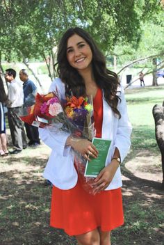 a woman in an orange dress is holding flowers and a book while standing under a tree