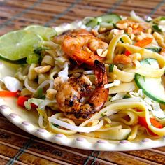 a white plate topped with noodles and shrimp next to cucumber wedges on a bamboo mat