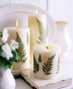 three white candles with green leaves on them sitting next to a plate and vase filled with flowers