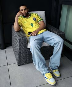 a man sitting in a chair wearing a yellow shirt and blue jeans with his hand on his chin