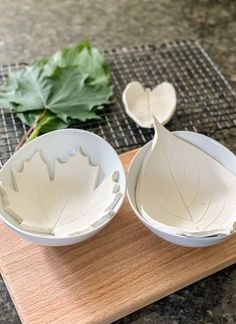 two white bowls sitting on top of a wooden cutting board next to leaf shaped spoons