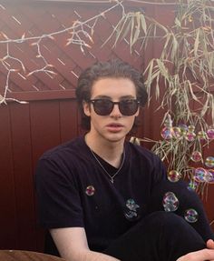 a man wearing sunglasses sitting in front of a tree with soap bubbles hanging from it