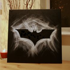 a chalk drawing of a batman symbol on a black canvas next to a lit candle