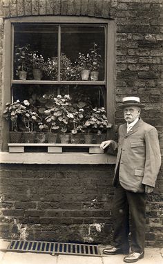 an old man standing in front of a window filled with potted plants and flowers