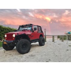 a red jeep parked on top of a sandy beach next to the ocean at sunset