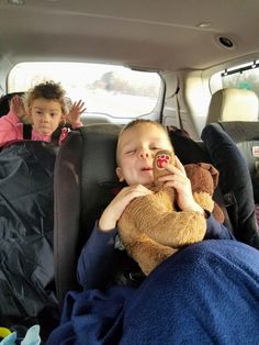 two children sitting in the back seat of a car, one holding a teddy bear