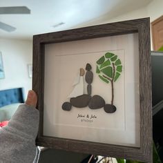 a person holding up a framed artwork with a tree and rocks in the shape of a couple