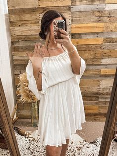 a woman taking a selfie in front of a mirror wearing a white dress and sandals