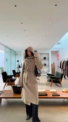 a woman taking a selfie with her cell phone in a clothing store while wearing a trench coat