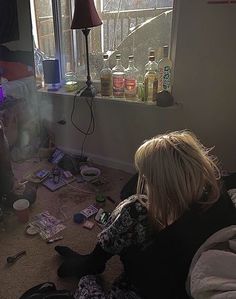 a woman sitting on the floor in front of a window with liquor bottles scattered around her